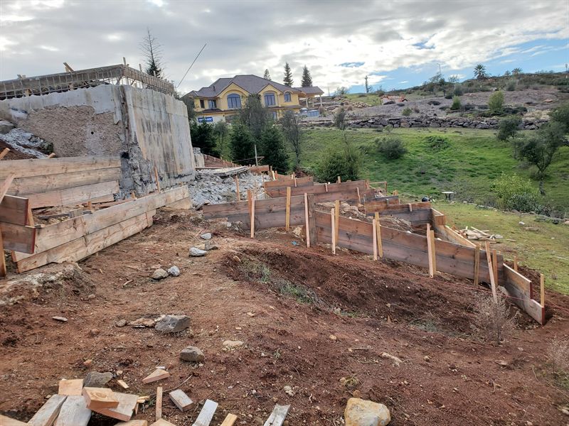 Concrete stem walls on a cleared hillside lot with standing houses on the ridge behind